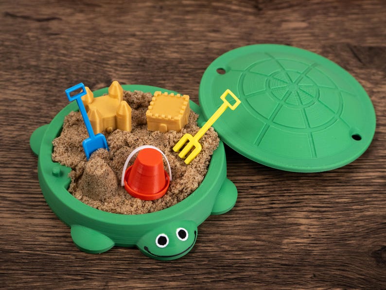 May include: A green turtle-shaped sandbox toy filled with sand and miniature toys. Includes a yellow rake, blue shovel, orange bucket, and yellow sandcastle and block. The lid is off to the side.
