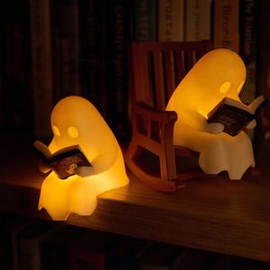 Two glowing reading ghost figurines on a wooden bookshelf: one perched on the shelf edge and one seated in a brown miniature rocking chair, each holding an open book.