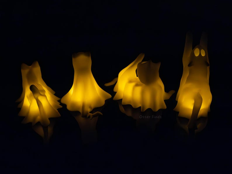 Pode incluir: Quatro pequenas figuras transl&uacute;cidas em forma de fantasma que brilham em amarelo, dispostas em fila. Cada figura apresenta uma forma suave e drapeada com detalhes faciais subtis, emitindo uma luz quente sobre um fundo escuro e refletor.