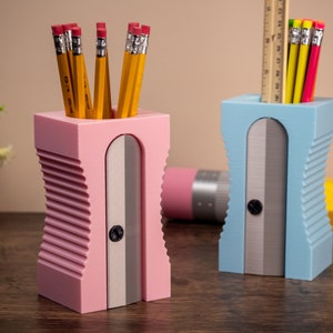 May include: Two pencil sharpeners, one pink and one blue, with pencil holders. The pink sharpener has a silver blade and is filled with yellow pencils. The blue sharpener has a silver blade and is filled with pencils and a ruler.