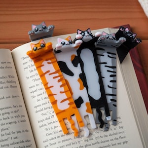 May include: A selection of cat-shaped bookmarks in different colours, such as orange, white, black, and grey, are positioned on the pages of an open book. The bookmarks have a cat design and are made of a glossy material.