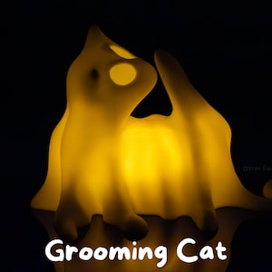 Pode incluir: Uma l&acirc;mpada amarela brilhante em forma de gato numa pose de limpeza contra um fundo preto. A l&acirc;mpada emite uma luz quente, destacando a silhueta felina e o texto Grooming Cat na parte inferior. O design apresenta bordas suaves e arredondadas.