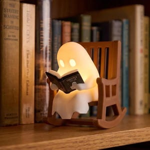 Glowing white reading ghost seated in a brown miniature rocking chair on a wooden bookshelf, holding an open black book with upright books behind it.