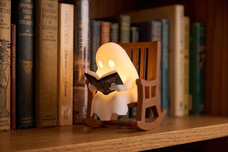 Glowing white reading ghost seated in a brown miniature rocking chair on a wooden bookshelf, holding an open black book with upright books behind it.