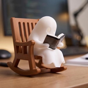 Unlit white reading ghost holding an open black book in a brown miniature rocking chair on a wooden desk, with a monitor, papers and lamp in the background.