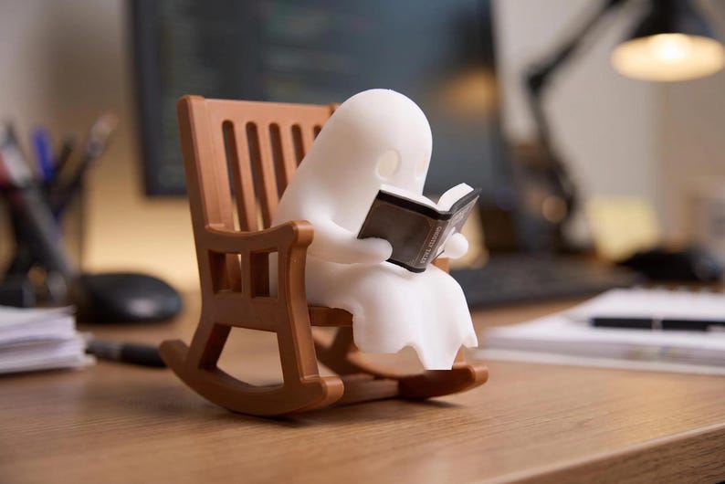 Unlit white reading ghost holding an open black book in a brown miniature rocking chair on a wooden desk, with a monitor, papers and lamp in the background.