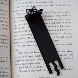 May include: Black cat bookmark with a cute face and paws, made of a ribbon-like material. The bookmark is placed on the pages of a book. The cat's face has pink accents.