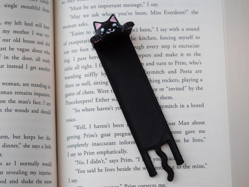 May include: Black cat bookmark with a cute face and paws, made of a ribbon-like material. The bookmark is placed on the pages of a book. The cat's face has pink accents.