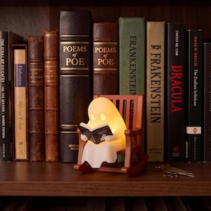 Wide bookshelf view showing the small glowing reading ghost in its brown rocking chair in front of full-size books, including Frankenstein and Dracula, illustrating the scale of the figurine.