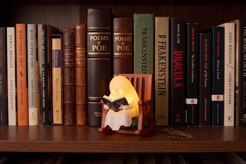 Wide bookshelf view showing the small glowing reading ghost in its brown rocking chair in front of full-size books, including Frankenstein and Dracula, illustrating the scale of the figurine.