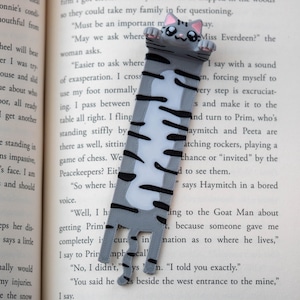 May include: A grey and white cat bookmark with black stripes and pink details. This bookmark is designed to be placed in a book. The cat's paws extend over the top of the page. The bookmark is made of a durable material.