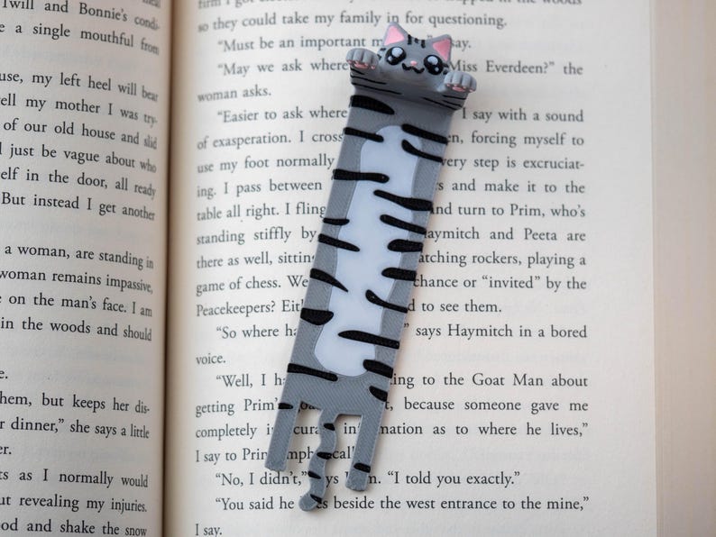 May include: A grey and white cat bookmark with black stripes and pink details. This bookmark is designed to be placed in a book. The cat's paws extend over the top of the page. The bookmark is made of a durable material.