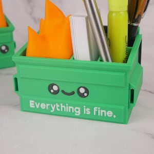 Puede incluir: Organizador de escritorio de pl&aacute;stico verde con forma de contenedor de basura con una cara sonriente y el texto "Todo est&aacute; bien". El organizador tiene una llama en la parte superior y contiene bol&iacute;grafos, l&aacute;pices y un resaltador.