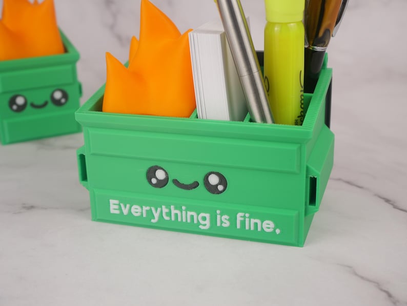 Puede incluir: Organizador de escritorio de pl&aacute;stico verde con forma de contenedor de basura con una cara sonriente y el texto "Todo est&aacute; bien". El organizador tiene una llama en la parte superior y contiene bol&iacute;grafos, l&aacute;pices y un resaltador.