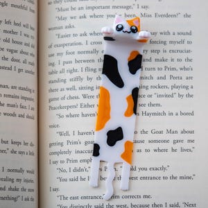 May include: A novelty cat bookmark, featuring a white body with black and orange markings. The bookmark is placed in a book, enhancing the reading experience. The cat has a cute, smiling face.