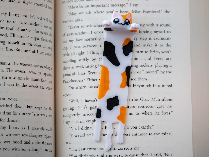 May include: A novelty cat bookmark, featuring a white body with black and orange markings. The bookmark is placed in a book, enhancing the reading experience. The cat has a cute, smiling face.