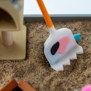 May include: A miniature cat litter box set with a white and orange shovel featuring a pink and black cow-like design. The set includes a tan cat tree and is placed on a bed of sand.