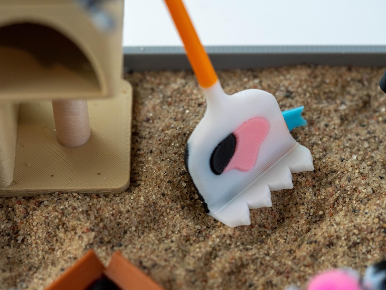 May include: A miniature cat litter box set with a white and orange shovel featuring a pink and black cow-like design. The set includes a tan cat tree and is placed on a bed of sand.