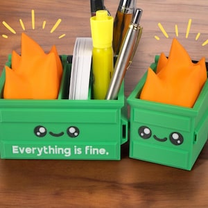 Puede incluir: Dos organizadores de escritorio verdes con forma de contenedor de basura con llamas naranjas en la parte superior. Los organizadores tienen una cara de dibujos animados linda con ojos negros y una sonrisa. El texto "Todo est&aacute; bien." est&aacute; impreso en la parte delantera del organizador.