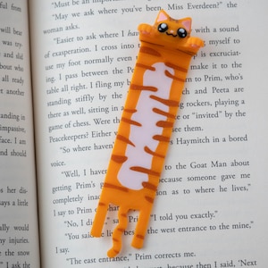 May include: A novelty bookmark in the shape of an orange and white cat. The bookmark is placed between the pages of a book. The cat has a smiling face and a long body with stripes.