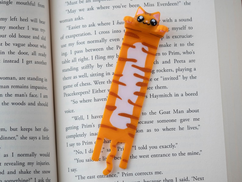 May include: A novelty bookmark in the shape of an orange and white cat. The bookmark is placed between the pages of a book. The cat has a smiling face and a long body with stripes.