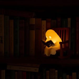 Wide view of a warmly glowing reading ghost in its brown rocking chair among upright books on a dark wooden bookcase, with another row of books below.