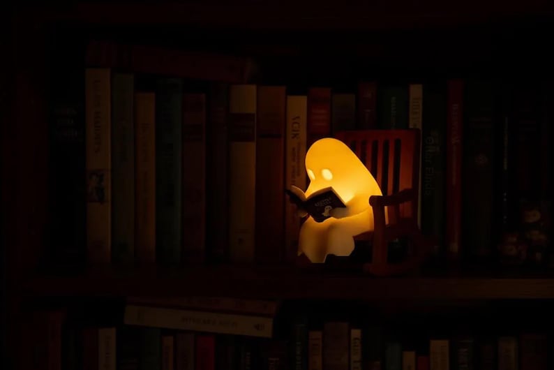 Wide view of a warmly glowing reading ghost in its brown rocking chair among upright books on a dark wooden bookcase, with another row of books below.