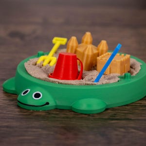 May include: A green turtle-shaped sandbox toy filled with sand, a yellow rake, a red bucket, a blue stick, and miniature sandcastles. The toy is designed for children and is set on a wooden surface.