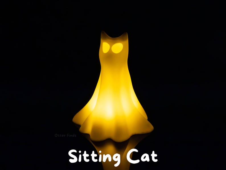 Pode incluir: Uma l&acirc;mpada amarela brilhante em forma de gato com uma silhueta suave e fluida que lembra um tecido drapeado. A l&acirc;mpada apresenta dois recortes distintos em forma de am&ecirc;ndoa para os olhos que emitem uma luz brilhante. O texto Sitting Cat est&aacute; impresso abaixo.