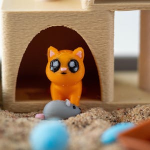 May include: A 3D-printed orange cat figurine with large black eyes stands in front of a tan cat house. A gray mouse figurine with a pink tail is in front of the cat. The scene is set in a sandbox with a blue ball and an orange scoop.