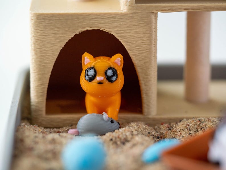 May include: A 3D-printed orange cat figurine with large black eyes stands in front of a tan cat house. A gray mouse figurine with a pink tail is in front of the cat. The scene is set in a sandbox with a blue ball and an orange scoop.