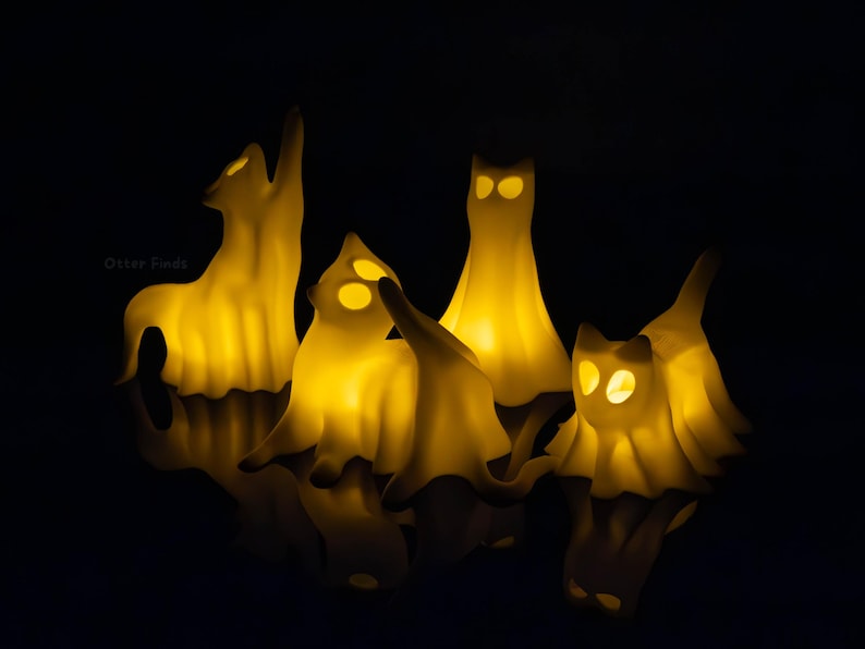 Pode incluir: Quatro pequenas figuras em forma de gato, na cor branco-sujo, iluminadas por dentro com uma luz amarela quente. Cada figura apresenta olhos recortados e uma apar&ecirc;ncia drapeada semelhante a um fantasma. O texto "Otter Finds" &eacute; vis&iacute;vel.