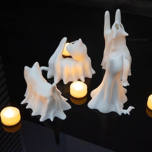 Pode incluir: Tr&ecirc;s figuras de gato em forma de fantasma branco com superf&iacute;cies lisas e mate. Cada gato &eacute; representado numa pose diferente, com um sentado, um de p&eacute; e um a esticar-se para cima. Tr&ecirc;s pequenas velas de ch&aacute; redondas emitem luz quente.