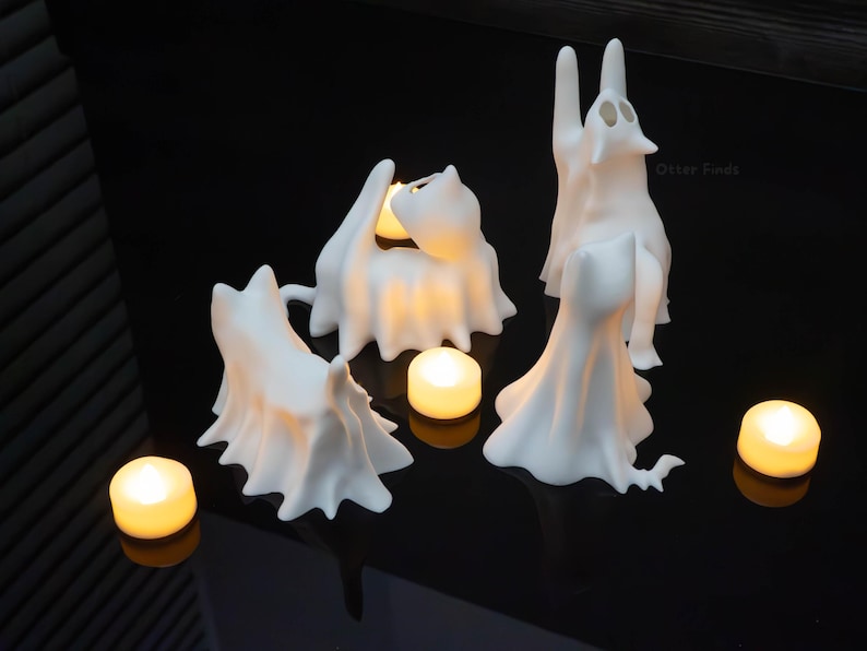 Pode incluir: Tr&ecirc;s figuras de gato em forma de fantasma branco com superf&iacute;cies lisas e mate. Cada gato &eacute; representado numa pose diferente, com um sentado, um de p&eacute; e um a esticar-se para cima. Tr&ecirc;s pequenas velas de ch&aacute; redondas emitem luz quente.