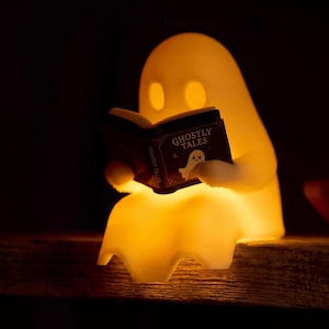 Close-up of a reading ghost glowing warm yellow on the edge of a wooden shelf, holding a black book titled Ghostly Tales; its draped sheet hangs over the edge.