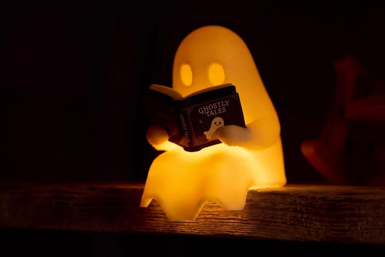 Close-up of a reading ghost glowing warm yellow on the edge of a wooden shelf, holding a black book titled Ghostly Tales; its draped sheet hangs over the edge.