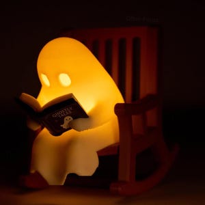 Close-up of a glowing reading ghost holding its Ghostly Tales book in a brown miniature rocking chair, photographed against a dark background.