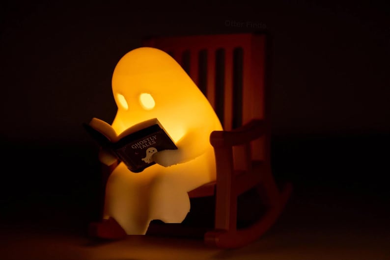Close-up of a glowing reading ghost holding its Ghostly Tales book in a brown miniature rocking chair, photographed against a dark background.