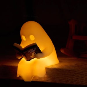 Glowing reading ghost perched on the edge of a wooden bookshelf, holding its open black book, with the empty brown rocking chair positioned behind it to the right.