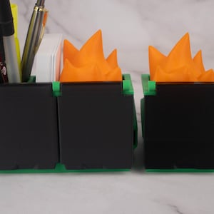 Puede incluir: Un organizador de escritorio negro con dos compartimentos, cada uno con una parte superior en forma de llama de color naranja brillante. El organizador est&aacute; hecho de pl&aacute;stico y tiene una base verde.