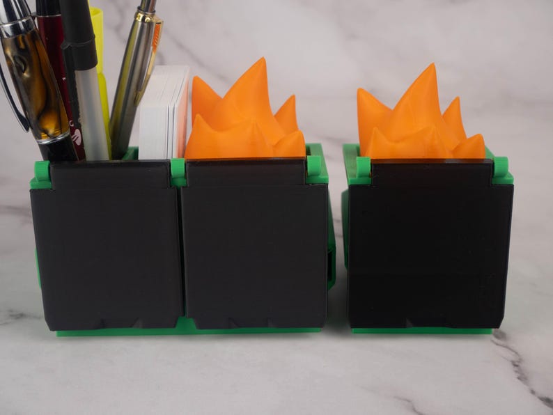 Puede incluir: Un organizador de escritorio negro con dos compartimentos, cada uno con una parte superior en forma de llama de color naranja brillante. El organizador est&aacute; hecho de pl&aacute;stico y tiene una base verde.