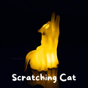 Pode incluir: Uma l&acirc;mpada amarela brilhante em forma de gato com um corpo transl&uacute;cido, representada numa posi&ccedil;&atilde;o sentada com a cabe&ccedil;a inclinada para cima. A luz emite um brilho quente e suave contra um fundo escuro. O texto "Scratching Cat" est&aacute; impresso em branco.