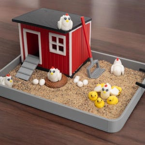 Puede incluir: Un set de jard&iacute;n zen en miniatura con tem&aacute;tica de granja que incluye un gallinero rojo y blanco, un rastrillo gris y varias figuras peque&ntilde;as de gallinas blancas, pollitos amarillos y huevos blancos en una bandeja rectangular gris con arena.