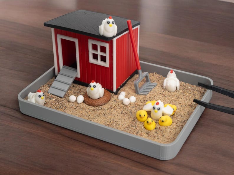 Puede incluir: Un set de jard&iacute;n zen en miniatura con tem&aacute;tica de granja que incluye un gallinero rojo y blanco, un rastrillo gris y varias figuras peque&ntilde;as de gallinas blancas, pollitos amarillos y huevos blancos en una bandeja rectangular gris con arena.