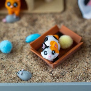 May include: A miniature cat figurine with black, white, and orange markings sits in a small brown box. A blue pom-pom, a yellow pom-pom, and a gray mouse figurine are scattered around the cat in a sand-filled container. Other cat-themed toys are in the background.