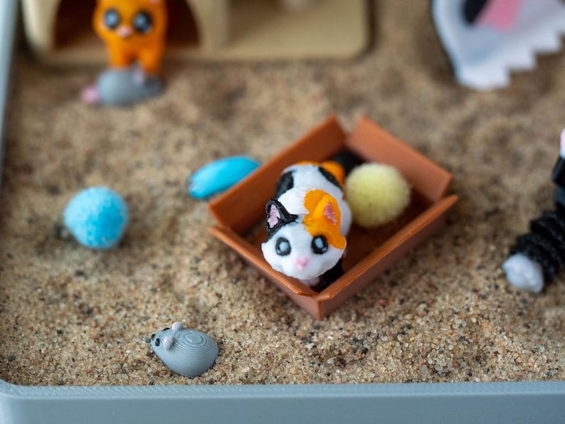 May include: A miniature cat figurine with black, white, and orange markings sits in a small brown box. A blue pom-pom, a yellow pom-pom, and a gray mouse figurine are scattered around the cat in a sand-filled container. Other cat-themed toys are in the background.