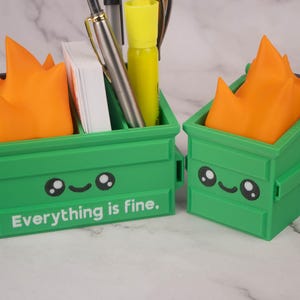 Puede incluir: Dos organizadores de escritorio de color verde en forma de contenedor de basura con llamas naranjas en la parte superior. El contenedor de basura de la izquierda tiene el texto "Todo est&aacute; bien." impreso en la parte delantera.