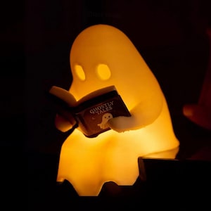 Close-up of the glowing ghost shelf sitter holding an open Ghostly Tales book, with its draped lower edge visible against a dark background and part of the chair at the right.