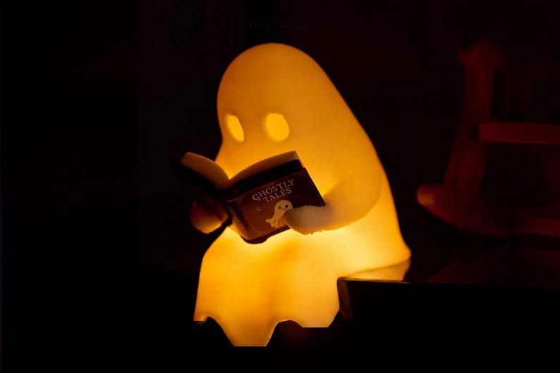 Close-up of the glowing ghost shelf sitter holding an open Ghostly Tales book, with its draped lower edge visible against a dark background and part of the chair at the right.