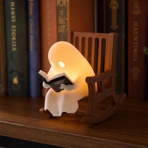 Three-quarter view of a softly glowing white reading ghost holding an open book in its brown rocking chair, displayed on a bookshelf beside a small potted plant.